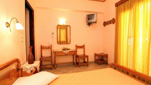 Et soveværelse med trémøbler, herunder en seng med gult sengetæppe, et skrivebord med spejl, stole og et lille fjernsyn på væggen. (AI-genereret tekst)