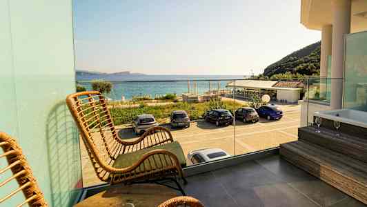 Udsigt fra altan med rottingstole og bord, glasrækværk, badekar på en platform samt biler parkeret ved stranden med havet i baggrunden. (AI-genereret tekst)