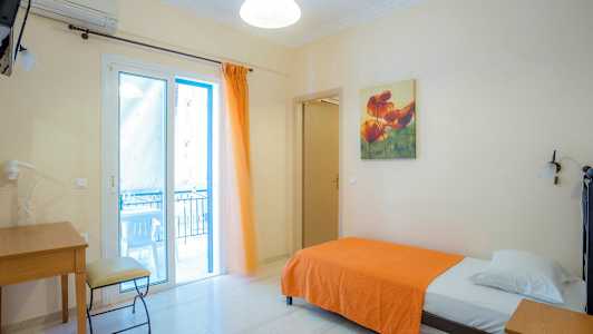 Et enkeltværelse med en seng med et orange tæppe, skrivebord og stol samt en glasdør, der fører til en balkon. (AI-genereret tekst)