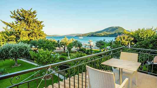 En balkon med bord og stole, udsigt over en grøn have, havet og bjerge i baggrunden. (AI-genereret tekst)