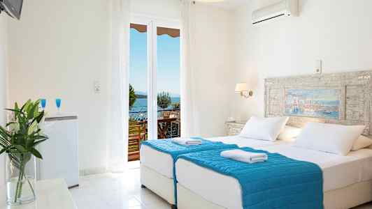 Et lyst hotelværelse med dobbeltseng og blåt sengetøj. Der er en balkon med udsigt over havet. (AI-genereret tekst)