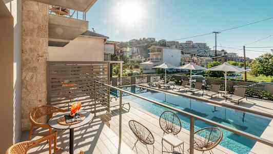 Billede af en solrig altan med bord og stole, et poolområde nedenfor med liggestole og parasoller. (AI-genereret tekst)