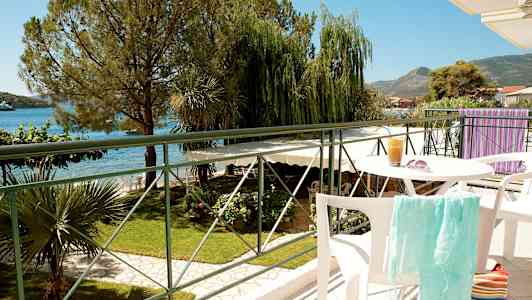 Udsigt fra en altan med bord og stole, med grønne områder, hav og bjerge i baggrunden. (AI-genereret tekst)