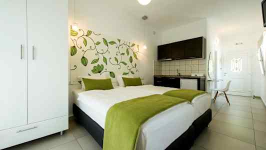 Et hotelværelse med en dobbeltseng dækket med hvidt sengetøj og grønne tæpper, et klædeskab, et tekøkken og en dør i baggrunden. (AI-genereret tekst)