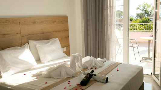 Hotelværelse med en dobbeltseng dekoreret med håndklæder og blomsterblade. En flaske vin og glas ligger på sengen. En balkon er synlig i baggrunden. (AI-genereret tekst)