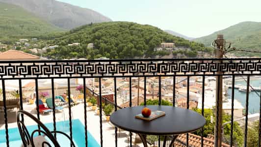 Udsigt over en balkon med et bord og dekorativt rækværk, poolområde, hustage og frodige bakker. (AI-genereret tekst)