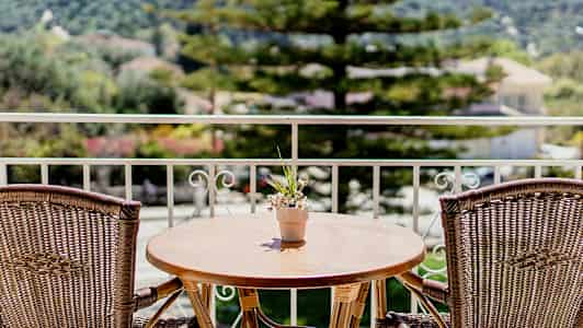 En altan med et træbord og to rattanstole samt en lille blomsterpotte med blomster på bordet. Træer og bygninger ses i baggrunden. (AI-genereret tekst)