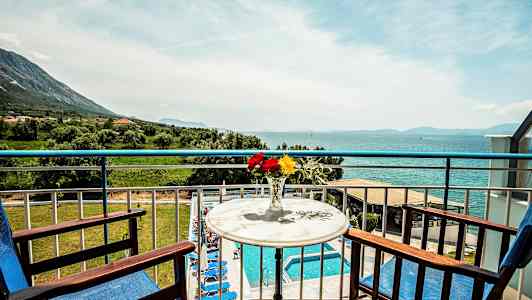 Udsigt fra en altan med to træstole og et bord med en vase med farverige blomster; i baggrunden ses en swimmingpool og et hav. (AI-genereret tekst)