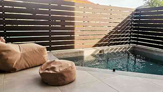 Lille pool med beige sækkestole på en stenoverflade, omgivet af et højt, mørkt hegn. (AI-genereret tekst)