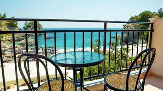 En balkon med et bord og to stole, med udsigt over havet og en strand. (AI-genereret tekst)