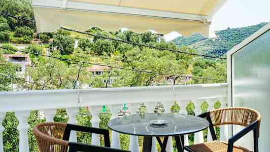 Udsigt fra en balkon med et lille rundt bord og to stole, omgivet af grønne områder og et bakket landskab. (AI-genereret tekst)