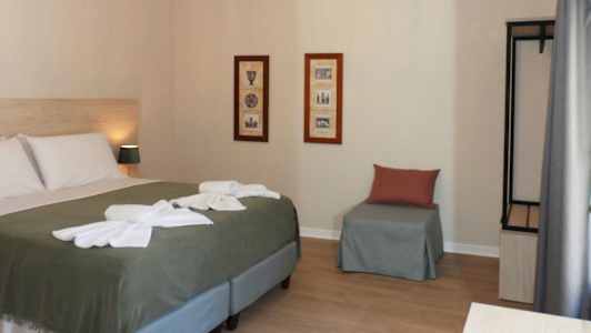 Et hotelværelse med en seng, to billeder på væggen, en stol med en pude og et åbent garderobeskab. (AI-genereret tekst)