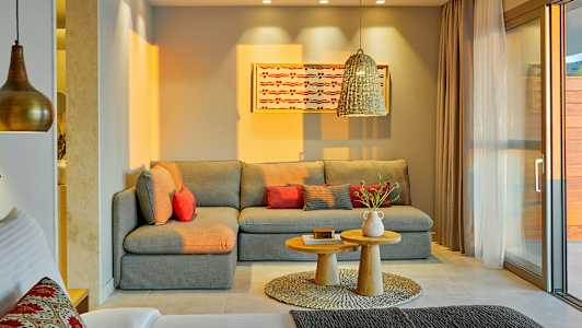 En stue med en grå hjørnesofa, rødlige puder, et par små træborde og et dekorativt tæppe. En hængende lampe og et maleri på væggen ses også. (AI-genereret tekst)