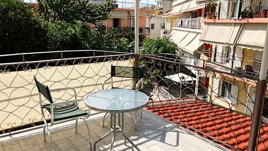 Altan med glasbord og to metalstole, med udsigt til grønne områder og omkringliggende bygninger. (AI-genereret tekst)