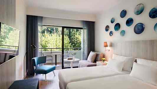 Moderne indrettet hotelværelse med to senge, en siddegruppe og balkon med udsigt til grønne områder. (AI-genereret tekst)