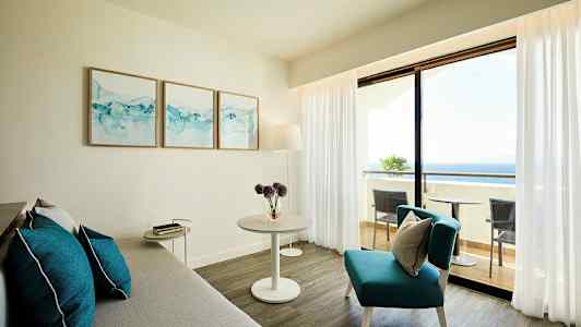 Lyst stue med grå sofa, blå dekorationspuder og bord, samt udsigt mod altan og hav. (AI-genereret tekst)