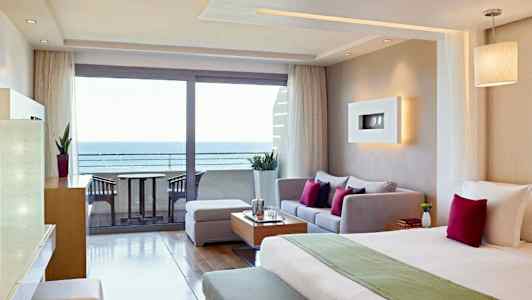 Et hotelværelse med moderne indretning, der inkluderer en seng, siddeområde og udsigt over havet gennem glasdøre. (AI-genereret tekst)