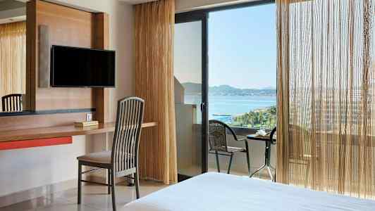 Et hotelværelse med skrivebord, stol og tv. Vindue og altan med havudsigt. (AI-genereret tekst)