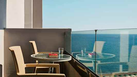 Udsigt fra en balkon med to stole og et bord med et glas vand og en bog foran havet. (AI-genereret tekst)