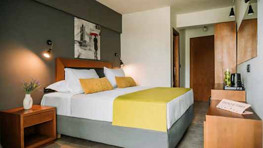 Et soveværelse med en dobbeltseng med hvidt og gult sengetøj, matchende puder, et natbord med en vase med blomster og et billede på væggen over sengen. (AI-genereret tekst)