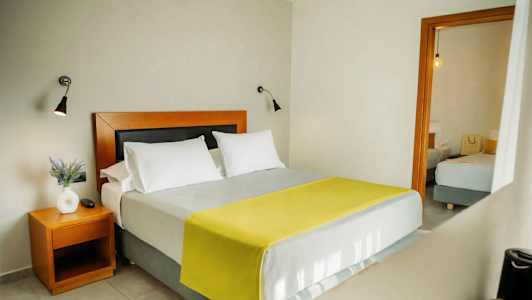 Soveværelse med en dobbeltseng, to hvide puder og et gult tæppe. Et sidebord med en hvid vase og en plante. (AI-genereret tekst)