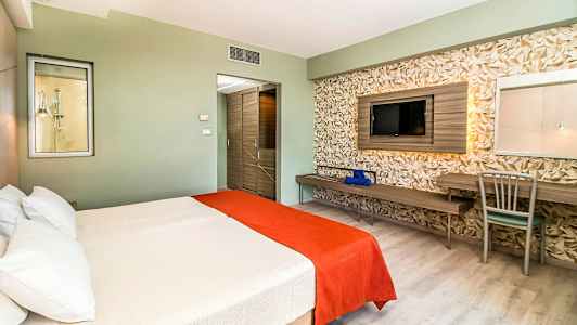 Hotelværelse med en dobbeltseng dækket med hvider og orange sengetæpper, en væg med geometrisk dekoration samt fjernsyn og skrivebord. (AI-genereret tekst)