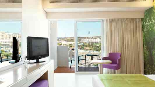 Hotelværelse med udsigt over havet, møbler inkluderer et TV, et skrivebord, en lilla stol og en stor seng. (AI-genereret tekst)