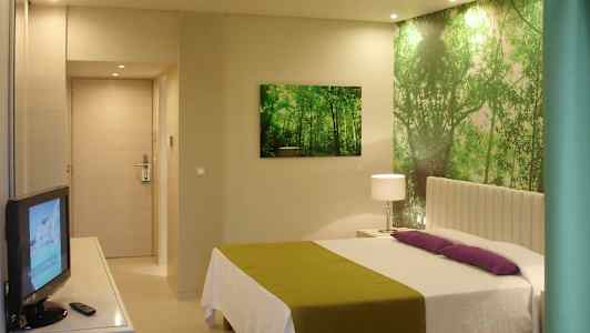 Et hotelværelse med en seng, grøn og hvid indretning, et tv og en vægdekoration af en skov. (AI-genereret tekst)