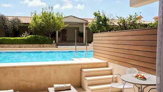 Udsigt fra en terrasse med liggestole og et bord med frugt, mod en pool og have. (AI-genereret tekst)