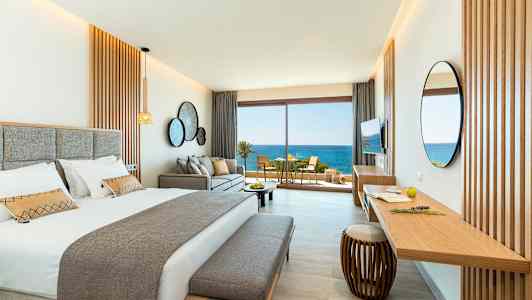 Et stilfuldt hotelværelse med en dobbeltseng, sofa og et skrivebord. Store glasdøre åbner ud til en altan med udsigt over havet. (AI-genereret tekst)