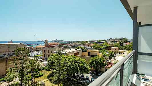 Udsigt fra en altan over en by med historiske bygninger, træer og en havn med både. (AI-genereret tekst)