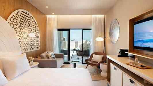 Et moderne hotelværelse med lyse møbler og dekorationer, herunder en seng, sofa, stol og TV. Værelset har store glasdøre, der fører ud til en balkon. (AI-genereret tekst)