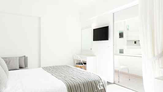 Interiøret i et lyst hotelværelse med en seng og skrivebord ved siden af en hvid stol. Et TV er monteret på væggen, og glasdøre fører til en balkon. (AI-genereret tekst)