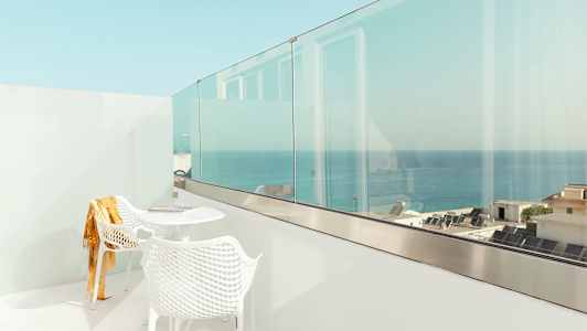 En hvid balkon med to stole og et bord, med udsigt over havet og bygninger. (AI-genereret tekst)