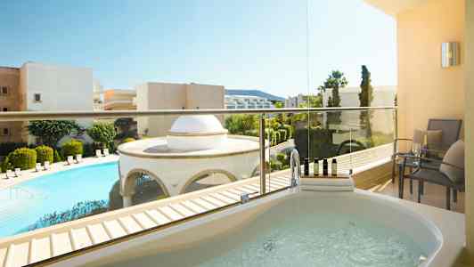 Billede af en altan med et spabad og siddepladser. Udsigt over en pool og omkringliggende bygninger. (AI-genereret tekst)