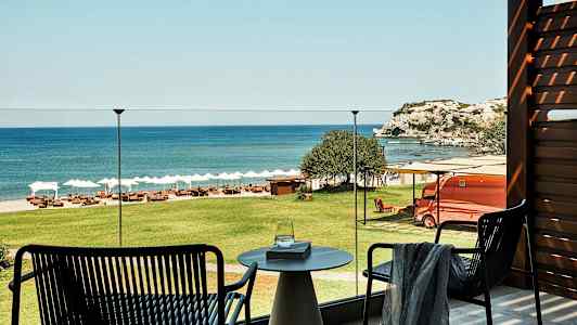 Udsigt fra en terrasse over grønt græs, en strand med liggestole og parasoller samt havet og klipper i baggrunden. (AI-genereret tekst)