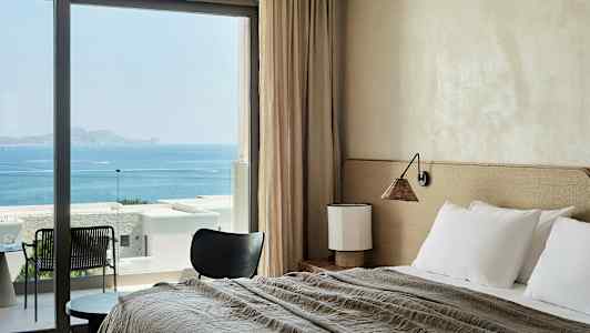 Et hotelværelse med seng, bord og stol, med havudsigt gennem store vinduer. (AI-genereret tekst)