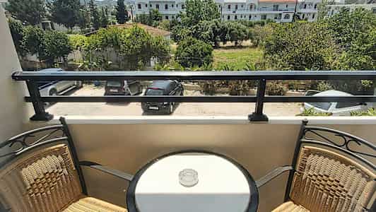 En altan med to stole og et rundt bord med et askebæger, med udsigt over en parkeringsplads og grønt område. (AI-genereret tekst)