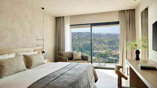 Et moderne hotelværelse med en stor seng, siddeområde og en balkon med udsigt over et grønt landskab. (AI-genereret tekst)