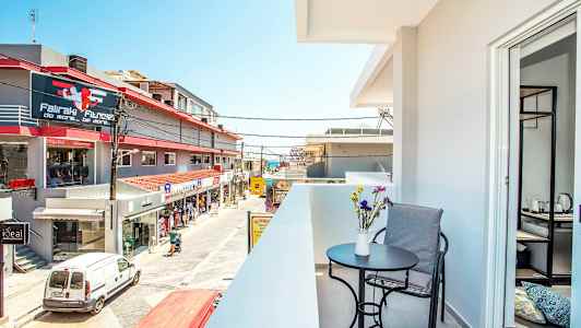 En altan med bord, stol og blomster i forgrunden samt udsigt over en gade med butikker og bygninger. (AI-genereret tekst)