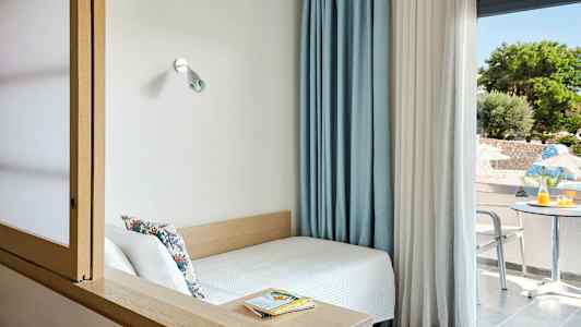 Et lille rum med en seng ved et vindue og udsigt til en terrasse med bord og stole. (AI-genereret tekst)