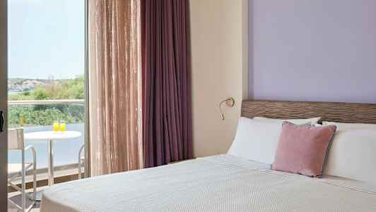 Et hotelværelse med en opredt seng, lilla vægge og gardiner samt en balkon med bord og to stole med udsigt til vegetation. (AI-genereret tekst)