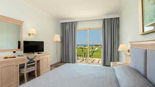 Et hotelværelse med en seng, skrivebord, tv og udsigt til en have og havet. (AI-genereret tekst)
