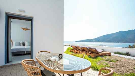 Udendørsmøbler med bord og stole foran en værelsesåbning og liggestole i en have med udsigt til havet. (AI-genereret tekst)