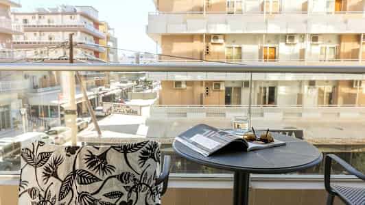 En altan med udsigt mod lejlighedsbygninger. På bordet ligger et glas, solbriller og et magasin. (AI-genereret tekst)