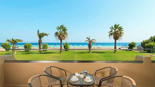 Udsigt fra en terrasse mod en strand med liggestole og palmer, med bord og stole i forgrunden. (AI-genereret tekst)