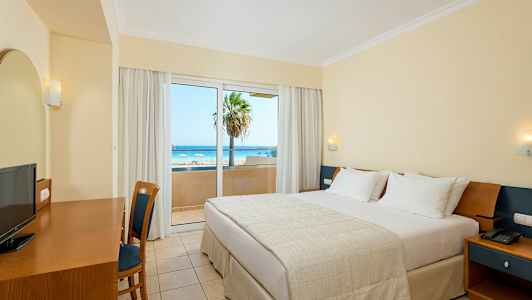 Hotelværelse med en dobbeltseng, skrivebord, stol og udsigt over en strand gennem en glasdør. (AI-genereret tekst)