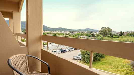 En altan med en stol og udsigt over en parkeringsplads samt grønne områder og bjerge i det fjerne. (AI-genereret tekst)