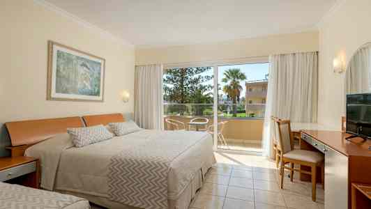 Et hotelværelse med redt seng, skrivebord og stole ved siden af en glasdør, der fører ud til en altan. (AI-genereret tekst)