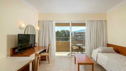 Et hotelværelse med et TV på et skrivebord, to enkeltsenge og en altan med havemøbler. (AI-genereret tekst)